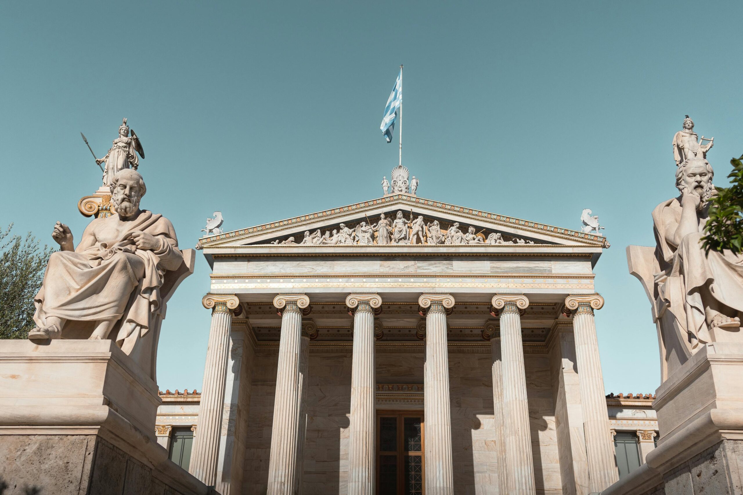 Classical architecture of the Academy of Athens with statues and Greek flag under a clear sky.